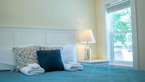A neatly made bed with a blue bedspread, two pillows, and a folded towel. There is a bedside lamp on a nightstand next to the bed. A window with blinds is visible in the background. - Beach Grass, 2 BR, South Haven, Walk to the Beach (South Haven)
