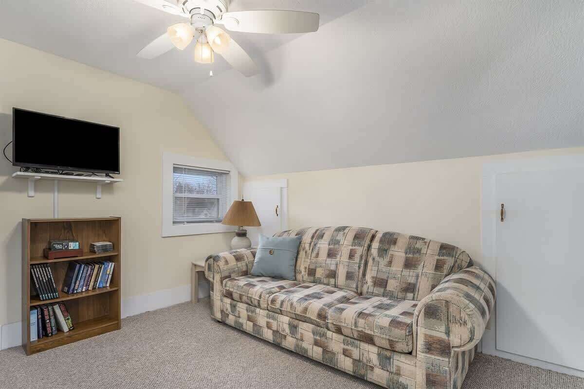 A cozy living room with a flat-screen TV, a bookshelf, a patterned sofa, a lamp, and a ceiling fan.