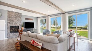 A modern living room with a stone fireplace, a large flat-screen TV, a grey sectional sofa, and a view of the outdoors through large windows.