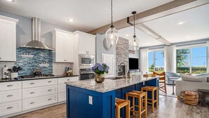 A modern kitchen with white cabinets, a blue island, wooden stools, and a stone backsplash.