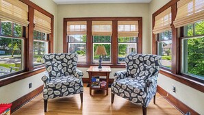 Une véranda avec deux fauteuils à motifs, une petite table avec une lampe et de grandes fenêtres équipées de stores.