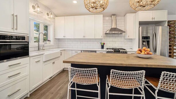 A modern kitchen with white cabinets, a central island, two wicker chairs, and pendant lighting. - SH Cottage (South Haven)