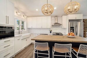 A modern kitchen with white cabinets, a central island, two wicker chairs, and pendant lighting. - SH Cottage (South Haven)