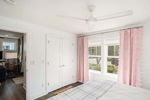 A bedroom with a bed, a ceiling fan, a window with pink curtains, and a view of the outdoors.