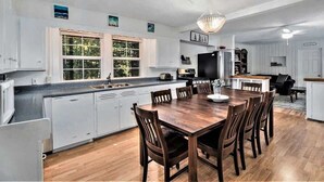 Una cocina amplia con una isla grande, una mesa de comedor de madera y sillas. Hay dos ventanas con marcos blancos, una araña y un refrigerador.