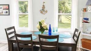 A dining area with a table, chairs, a vase with flowers, and a view of a grassy yard through large windows. - Mail Legacy Cottage in South Haven • Dogs Welcome (South Haven)