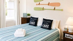 A bedroom with a bed, pillows, a nightstand, a lamp, and a window with curtains. - Mail Legacy Cottage in South Haven • Dogs Welcome (South Haven)