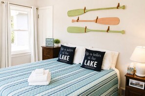 A bedroom with a bed, pillows, a nightstand, a lamp, and a window with curtains. - Mail Legacy Cottage in South Haven • Dogs Welcome (South Haven)