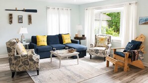 A living room with a blue sofa, patterned armchairs, a wooden chair, a coffee table, and a view of greenery outside. - Mail Legacy Cottage in South Haven • Dogs Welcome (South Haven)