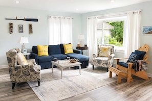 Un salon avec un canapé bleu, des fauteuils à motifs, une chaise en bois, une table basse et une vue sur la verdure à l’extérieur.