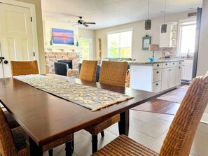 Uma área de refeições com mesa de madeira e cadeiras de vime, cozinha ao fundo e uma lareira.