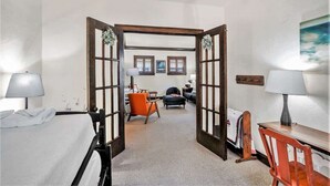 Ein Hotelzimmer mit einem Bett, einem Schreibtisch, einem Sessel und einer Couch. Es gibt zwei Fenster mit Vorhängen, eine Tür mit Holzrahmen und ein Bild an der Wand.