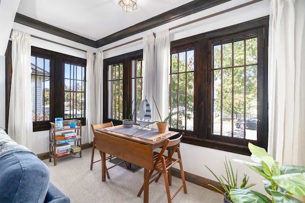 Ein Zimmer mit einem Holzesstisch, Stühlen, einem Bücherregal und großen Fenstern mit Vorhängen.