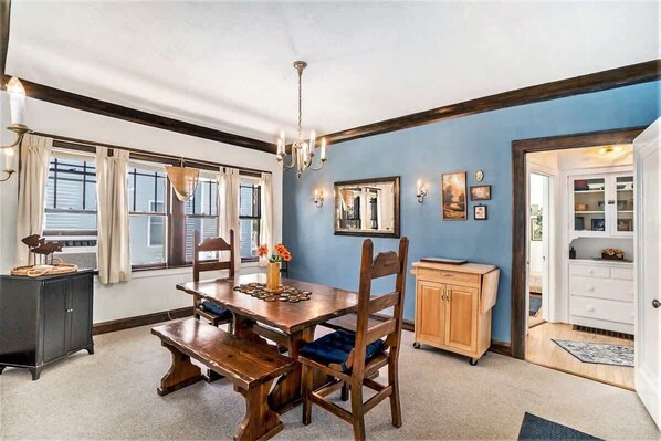 A dining room with a wooden table and chairs, a chandelier, and a view of the outdoors through large windows. - Breezy Beach Bungalow (South Haven)