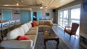 Un vaste espace de vie doté d’un plafond en bois, d’un grand canapé d’angle beige agrémenté de coussins rouges et jaunes, d’une table basse en bois et offrant une vue sur l’extérieur grâce à de grandes fenêtres.