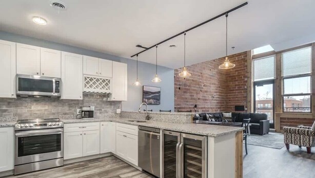 A modern kitchen with white cabinets, a built-in oven, a microwave, and a wine cooler. There is a dining area with a black sofa and a patterned armchair.