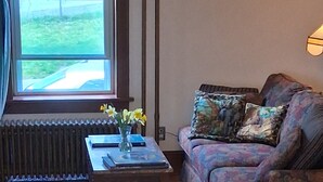 A living room with a sofa, a coffee table with a vase of flowers, a radiator, and a window with a view of the outside.