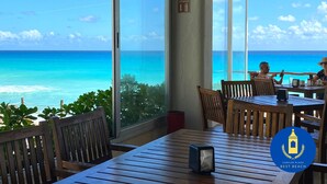 Un restaurante con mesas y sillas de madera, grandes ventanales con vista al océano y un letrero que dice 'Extinctor' en la pared.