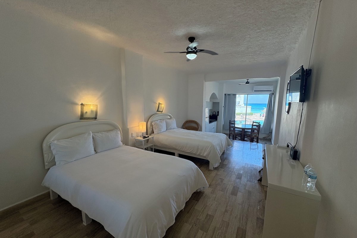 Une chambre d’hôtel avec deux lits, un ventilateur de plafond, une petite table avec une chaise et une vue sur la plage.