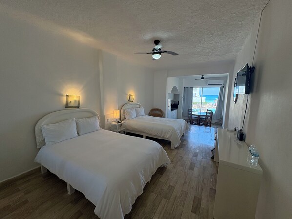 A hotel room with two beds, a ceiling fan, a small table with a chair, and a view of the beach.