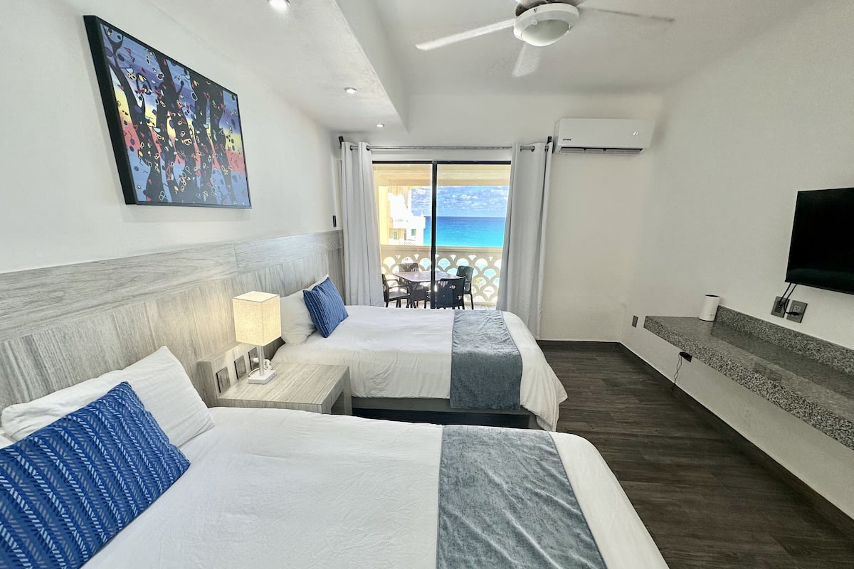 Une chambre d’hôtel avec deux lits, un ventilateur de plafond, une télévision et un balcon avec vue sur l’océan.