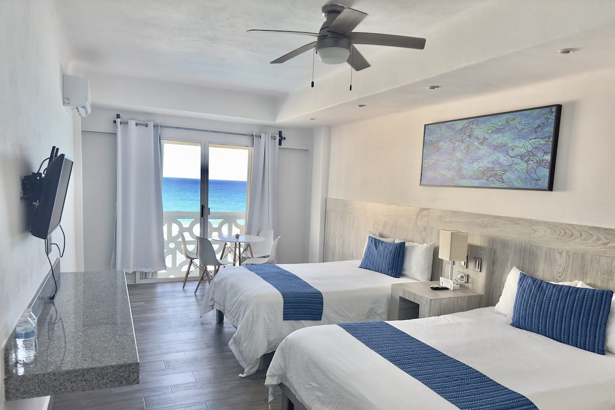 Une chambre d’hôtel avec deux lits, un bureau, une chaise, un ventilateur de plafond et une vue sur l’océan.