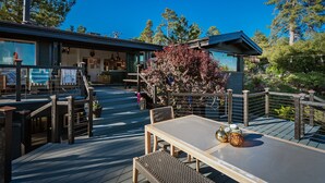 Un terrazzo in legno con tavolo e sedie, un piccolo giardino con un albero e un edificio con ampie finestre e un balcone.