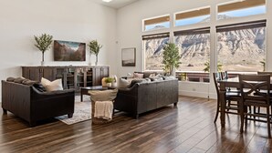 A spacious living room with a large sectional sofa, a dining area with wooden chairs, and a view of a mountainous landscape through large windows. - Large 9 Bedroom Home w/Private Pool, Indoor Sports Court & Arcade Room (Hurricane)