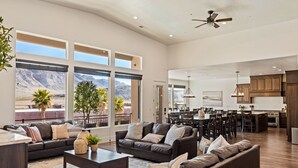 A spacious living room with a large window overlooking a mountainous landscape, a ceiling fan, and a modern kitchen in the background. - Large 9 Bedroom Home w/Private Pool, Indoor Sports Court & Arcade Room (Hurricane)