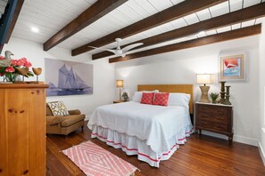 Une chambre avec un plafond en bois, un lit avec une literie blanche, une table de chevet, une lampe, une chaise en osier et un tableau représentant un voilier.