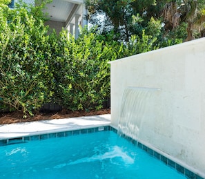 A swimming pool with a waterfall feature, surrounded by lush greenery and a white wall.