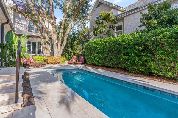 Une piscine dotée d’une terrasse carrelée, entourée d’une végétation luxuriante et d’une maison avec un balcon.