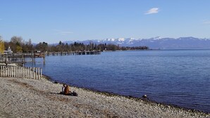 A lakeside view with a wooden dock, pebbled shore, and mountains in the background. - Manuele Ficano - Modern vacation apartment on beautiful Lake Constance (Kressbronn am Bodensee)