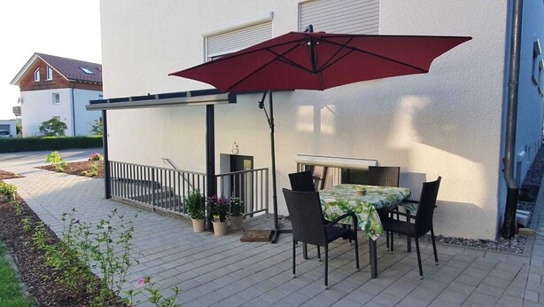 A patio with a table and chairs, a red umbrella, and potted plants. - Manuele Ficano - Modern vacation apartment on beautiful Lake Constance (Kressbronn am Bodensee)