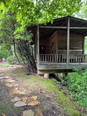 Un chalet rustique en bois, doté d’un porche, entouré d’une végétation luxuriante et accessible par un sentier pavé de pierres.