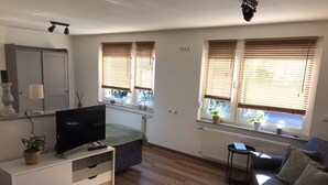 A modern living room with a flat-screen TV, a sofa, a dining table, and two windows with blinds.
