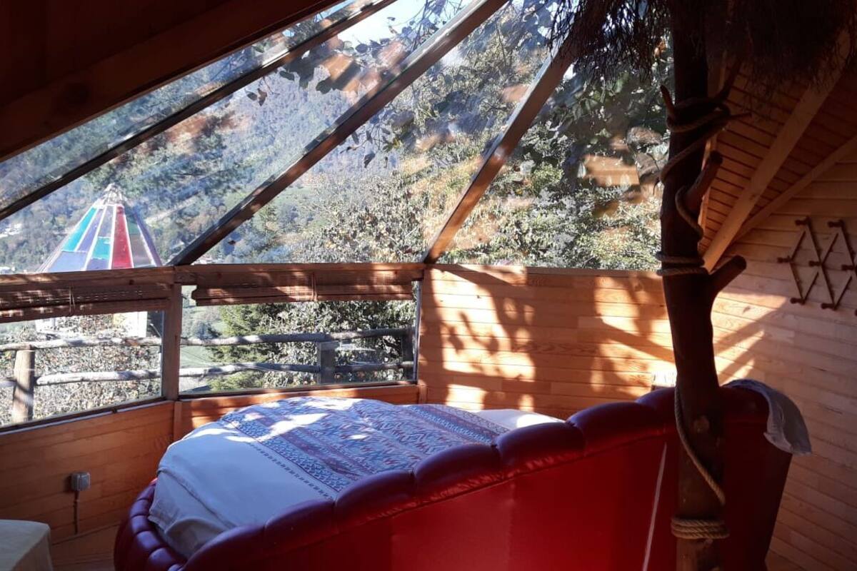 A wooden cabin with a sloped roof, a bed with a patterned blanket, and a view of a colorful structure in the distance.