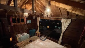 A cozy bedroom with a bed, a small table, and a window overlooking a cityscape at night. - Olimbera Köy Evi (Arhavi)