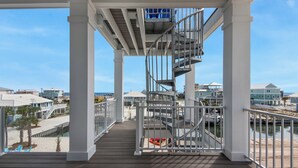 A wooden deck with a metal staircase leading to a covered area, overlooking a beach and residential buildings. - Blue Chair | 🚤 Boater’s Paradise | Pool | Dock | Outdoor Bar | Pet Friendly 🐾 (Navarre)