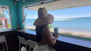 A person seated by a window with a view of the beach, holding a tumbler. - Unique and Private Panoramic Ocean View Penthouse in Cabo Rojo, Puerto Rico (Cabo Rojo)