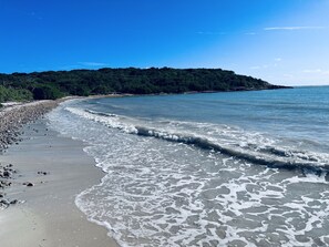 Una playa con agua azul cristalina, un litoral rocoso y una colina boscosa al fondo.