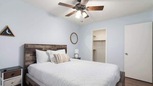 A bedroom with a bed, a nightstand, a ceiling fan, a framed picture, and a door.
