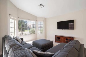 A living room with a sofa set, a TV mounted on the wall, and a window with blinds.