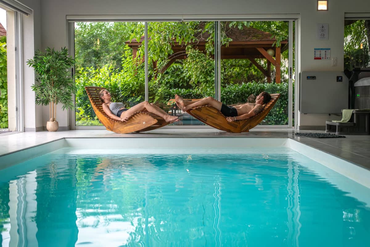 A pool area with two lounge chairs facing each other, a large window with a view of greenery, and a potted plant to the left.