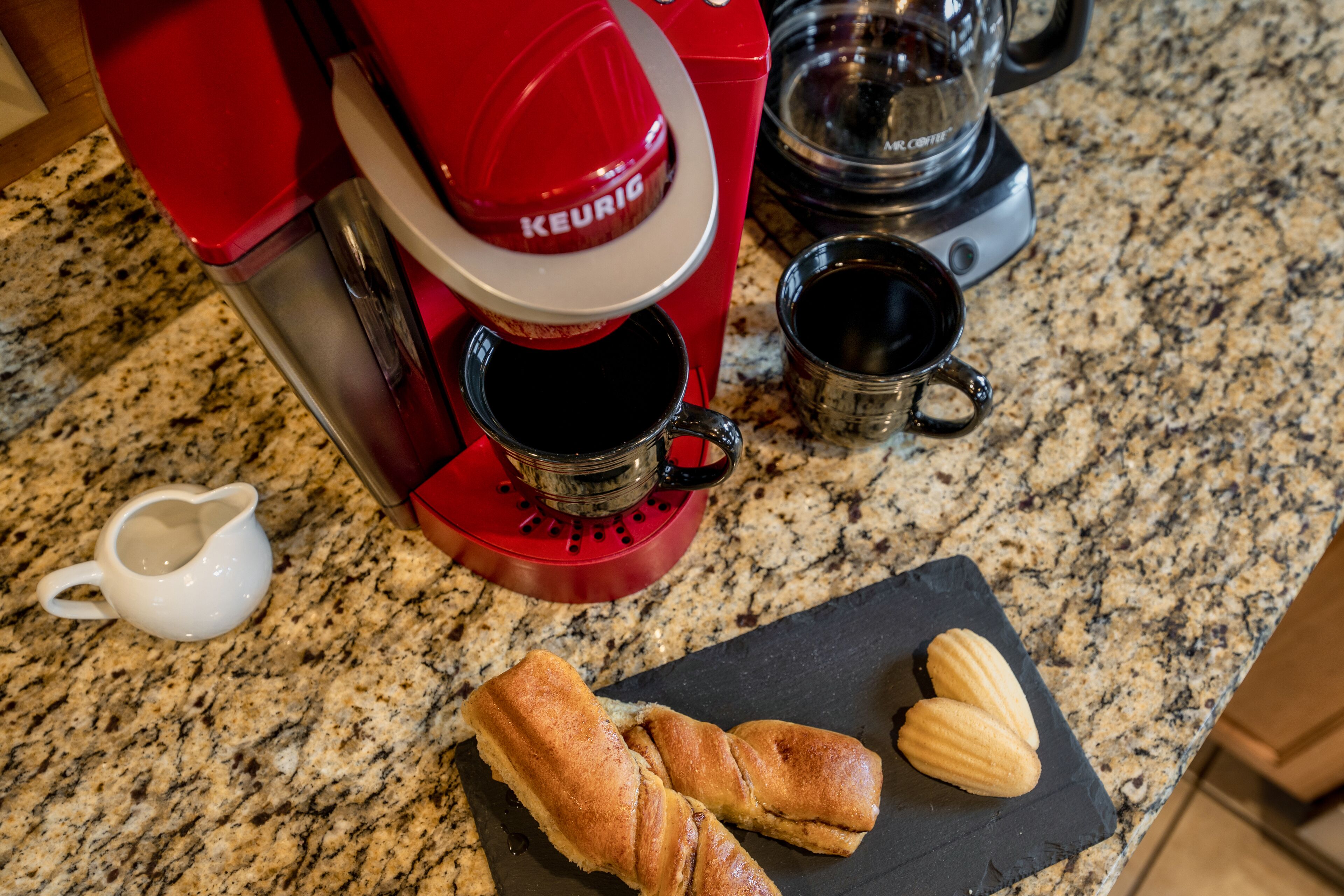 Une machine à café Keurig rouge est posée sur un comptoir en granit, accompagnée de deux tasses à café noires et d’un petit pichet blanc. Deux croissants se trouvent sur une assiette en ardoise noire.