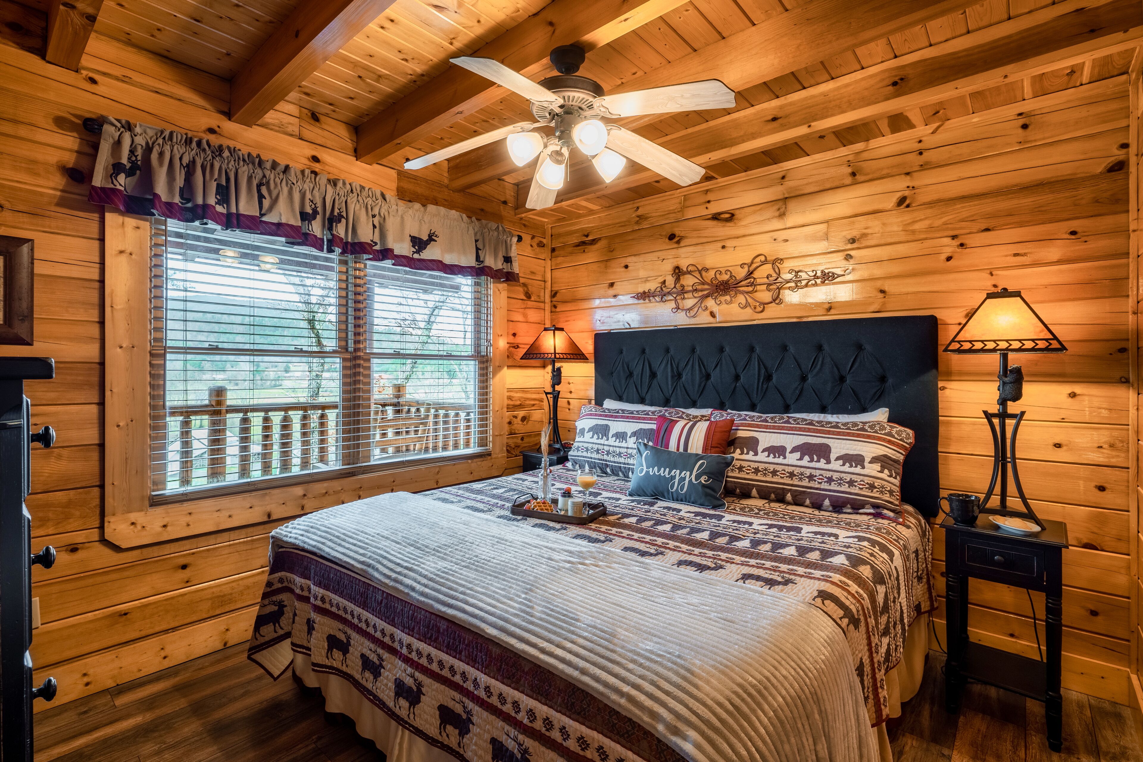 Ein Schlafzimmer einer Holzhütte mit einem Bett, einem Deckenventilator, einem Fenster mit Jalousien, einem Nachttisch und einer Lampe.