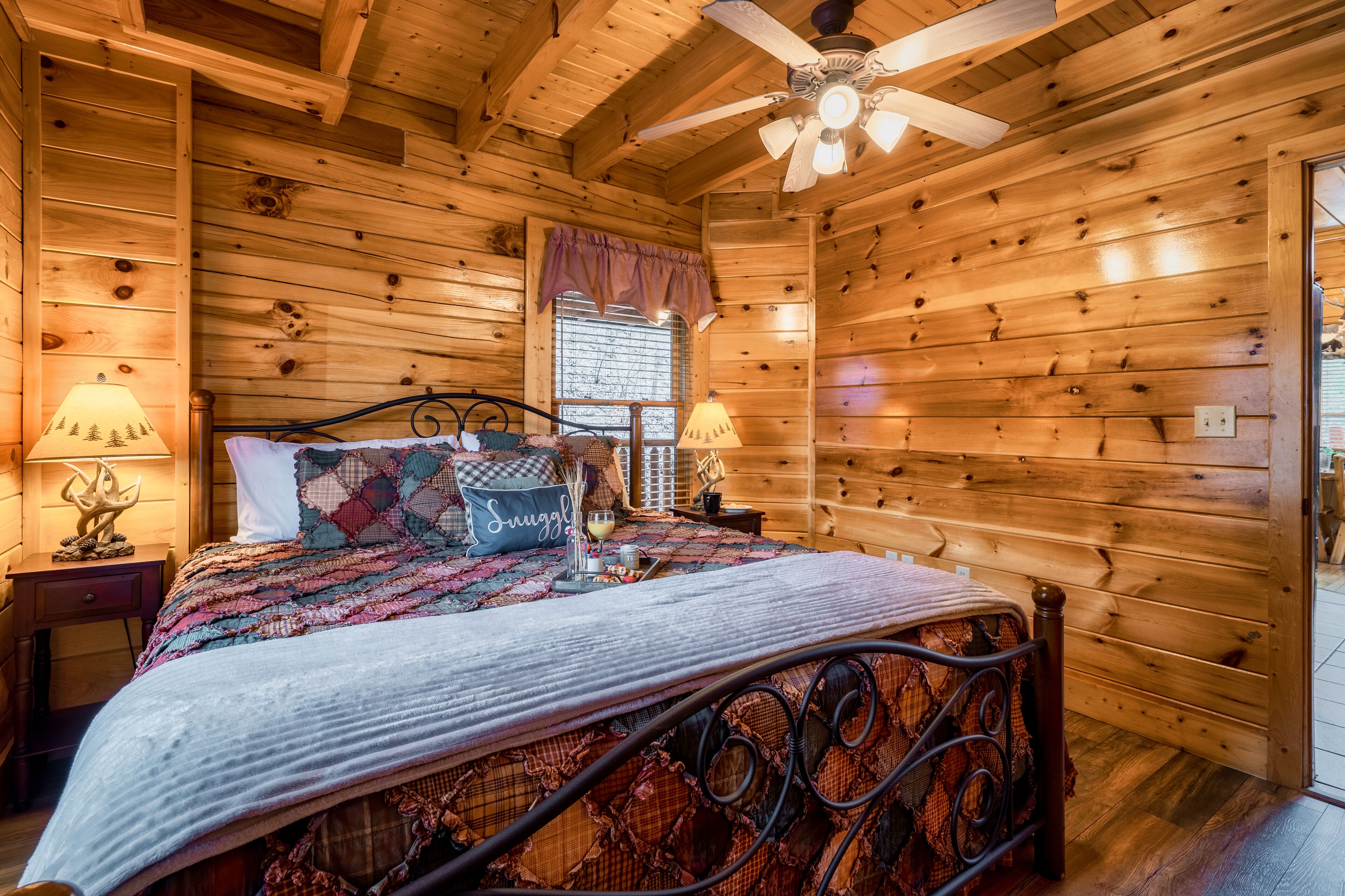 Camera da letto in una baita di legno, con un letto, due lampade ai comodini, una ventola a soffitto e una finestra con vista.
