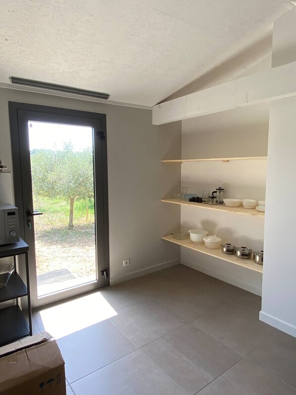 Ein Zimmer mit einer Schiebeglastür, die zu einem Garten führt, eingebauten Regalen mit Küchenutensilien und einem Fliesenboden.