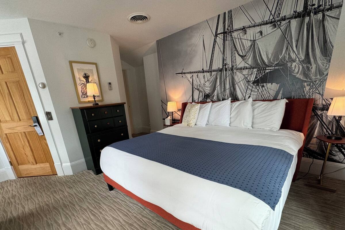 A neatly made bed with a blue and white bedspread, a wooden door, a dark-colored dresser, and a large wall tapestry depicting a ship.