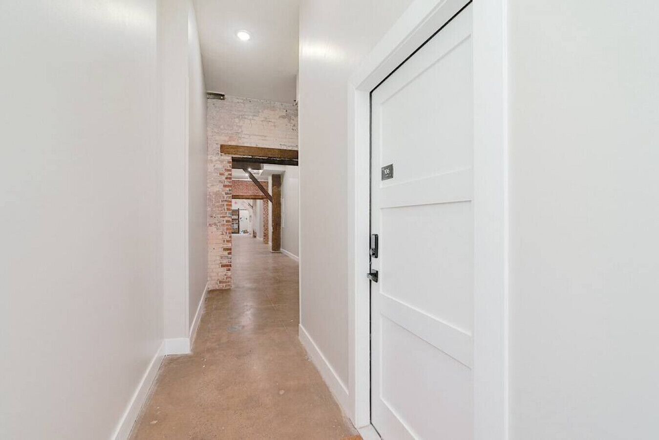 Un pasillo estrecho con una puerta blanca a la derecha, un muro de ladrillo a la izquierda y un techo con vigas de madera.